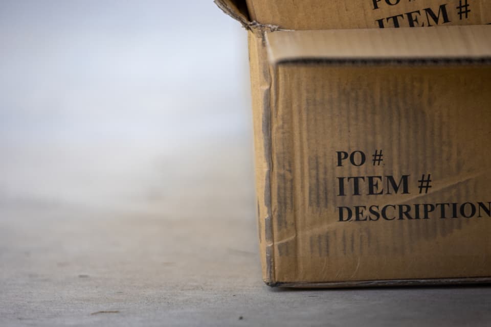 A close-up of a cardboard shipping box printed with 'PO#, Item#, Description'