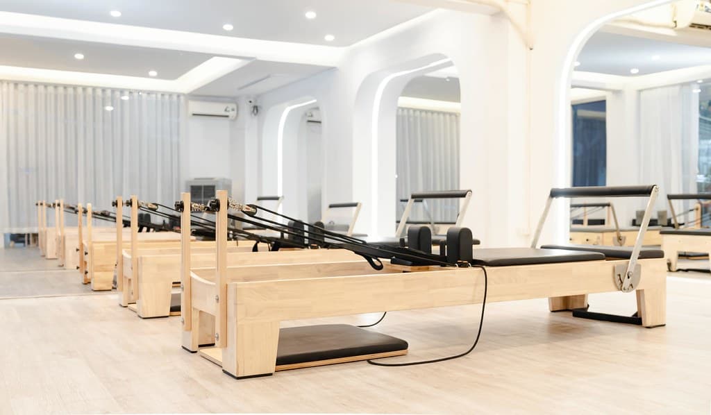 A bright studio with white walls and wide curtains, lined with wooden reformer pilates machines