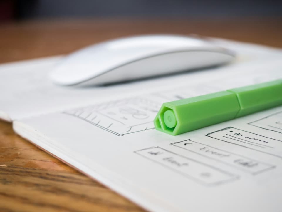 A green pen resting on a hand-drawn wireframe sketch in a notebook, with a blurred mouse in the background
